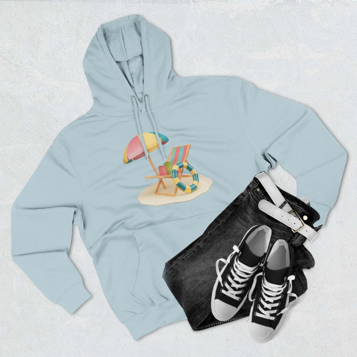 Light blue Sunny Resort Hoodie with beach chair design, paired with black jeans and sneakers on a textured background.